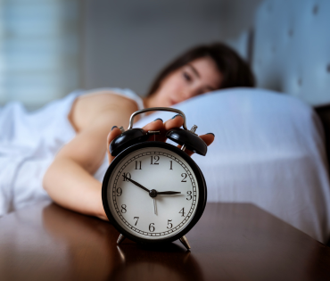 woman lying in bed reaching out to her alarm clock